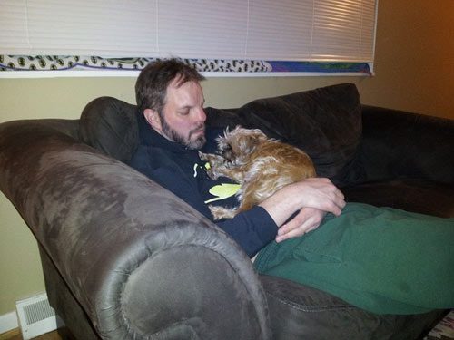A man sitting on top of a couch with his arm around a cat.